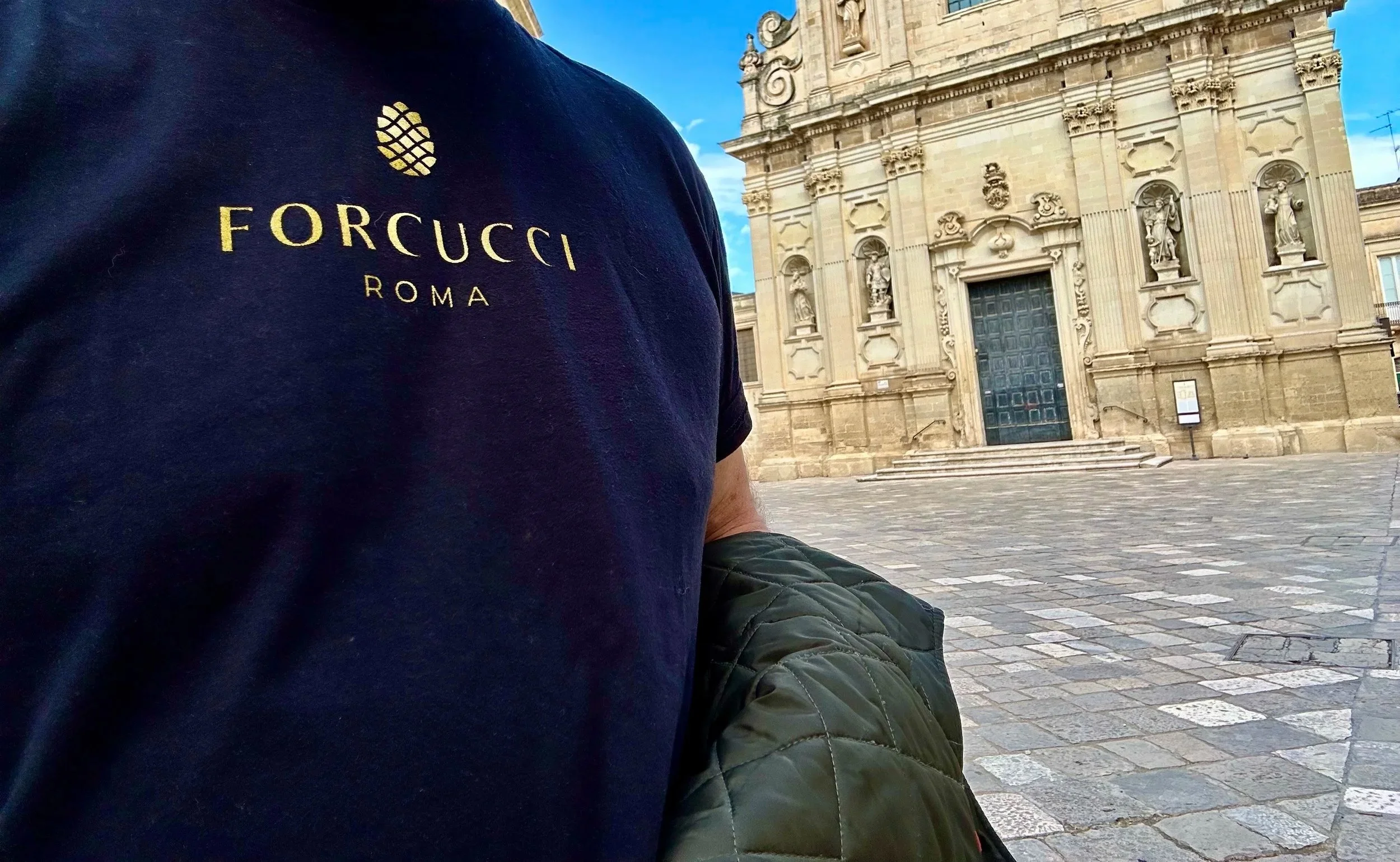 Person wearing a navy blue t-shirt with gold lettering that says 'FORCUCCI ROMA,' standing in front of an ornate stone building with statues and a large door, in a European city square.