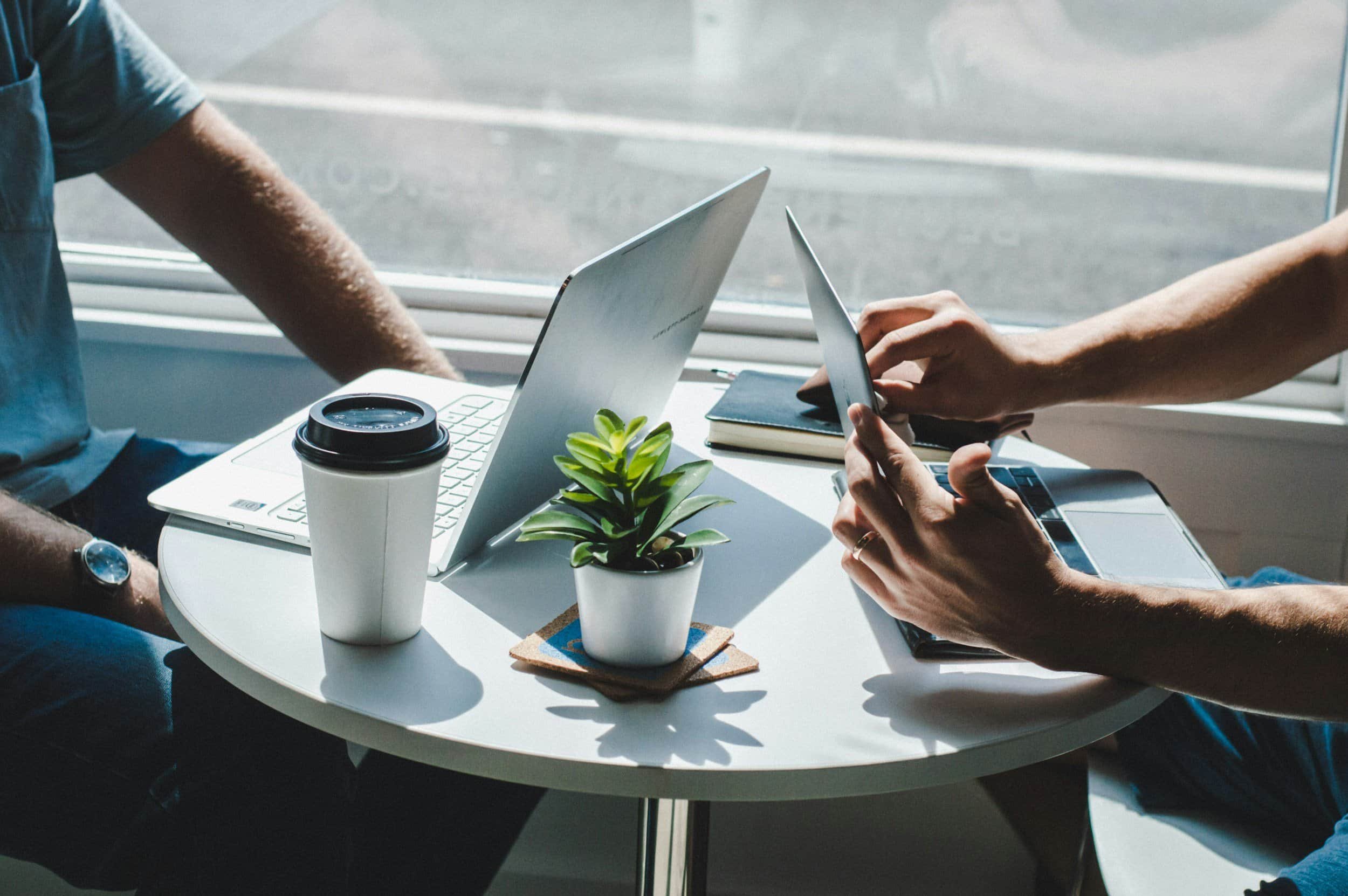 Two people sit at a round white table with a laptop, a plant, a coffee cup, a notebook, and a tablet on it, sunlight coming through a window.