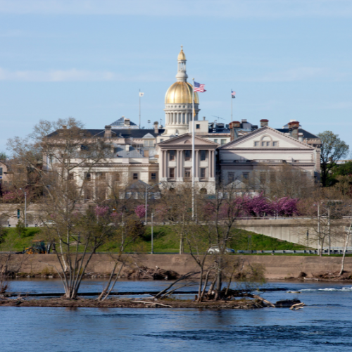 State capitol building with a gold dome beside a river, surrounded by trees and spring foliage under a clear sky.