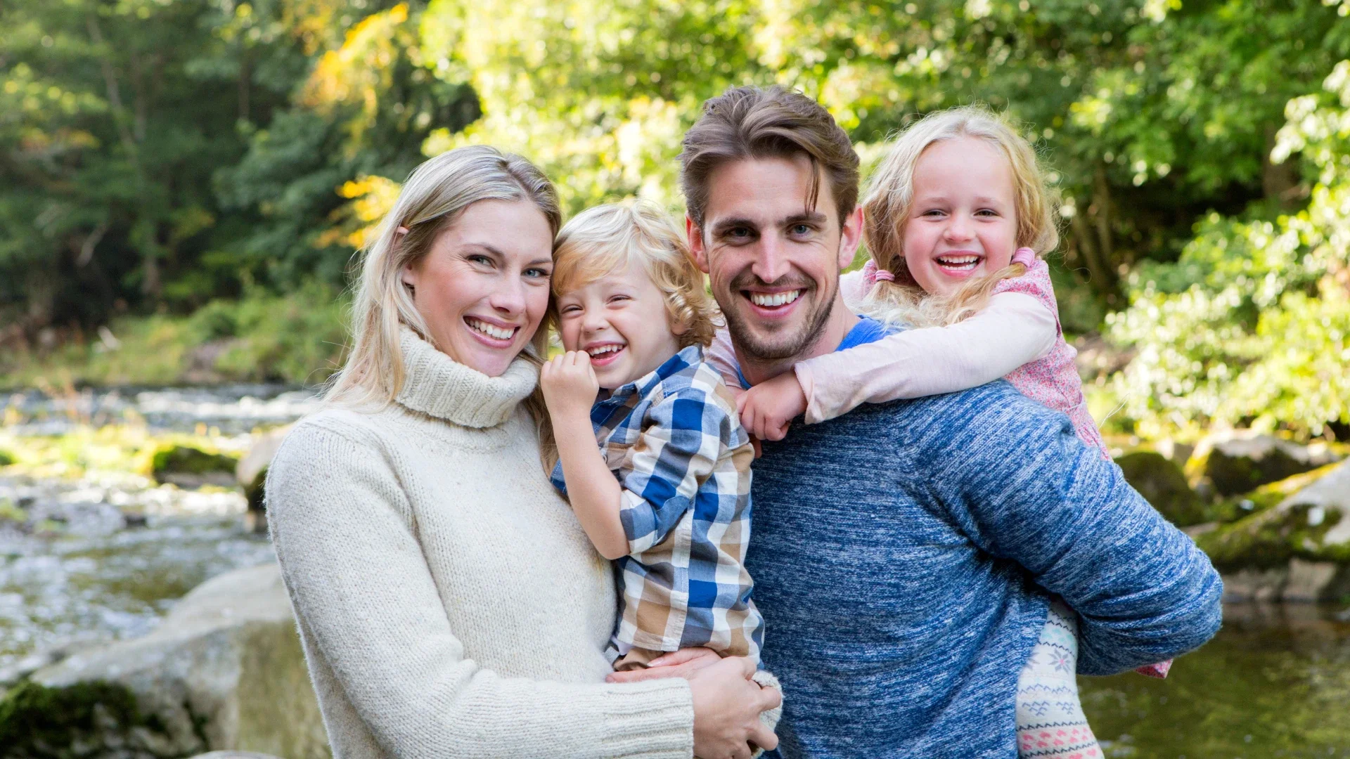 Happy family of four smiling together outdoors by a river, with parents holding their young children in a sunny natural setting.
