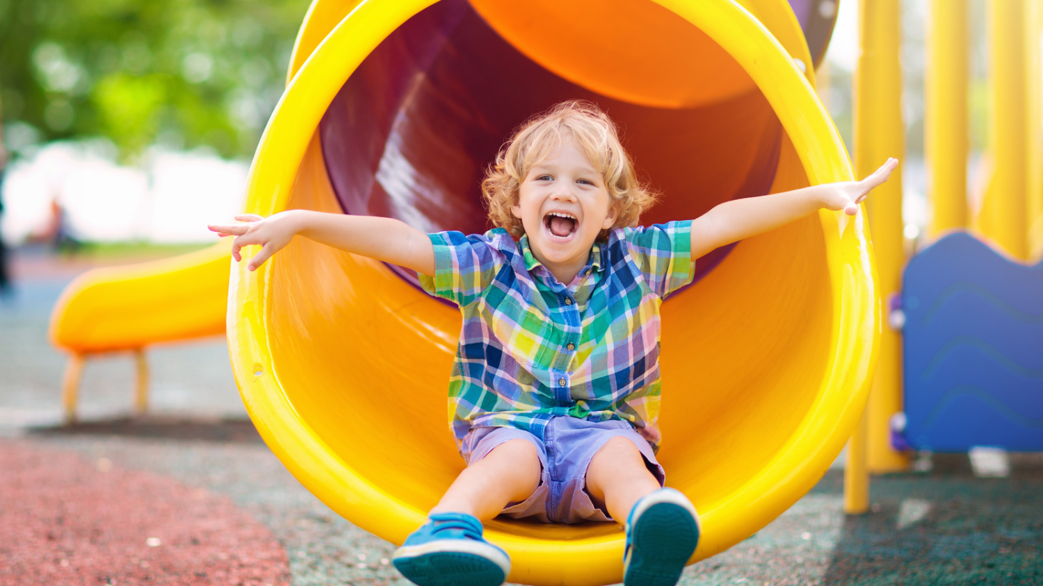 A smiling child with arms outstretched sitting on the end of a yellow slide in a NYC playground