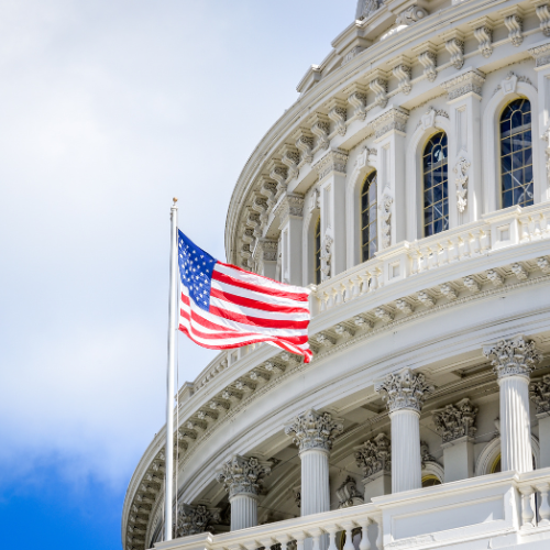 American flag flying in front of the U.S. Capitol building with classical columns and detailed architecture under a partly cloudy sky.