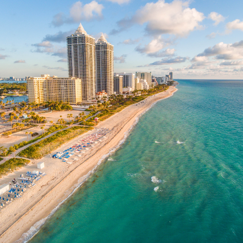 Aerial view of a sandy beachfront with turquoise ocean water and high-rise hotels lining the coast under a partly cloudy sky.