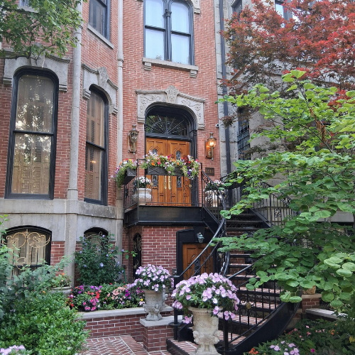 Brick townhouse facade with ornate doorway, flower-filled balcony, and staircase surrounded by lush greenery and garden plants.