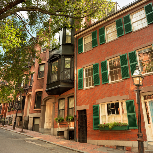 Historic brick townhouses with green shutters and bay windows along a quiet, tree-lined street.