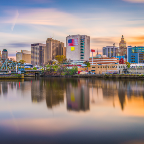City skyline reflected in a calm river at sunset, with mid-rise buildings and a large American flag mural on one facade.