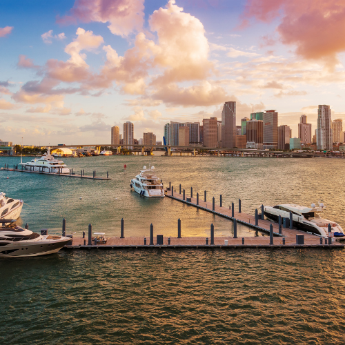 Boats docked at a marina with a city skyline in the background under a partly cloudy sunset sky.