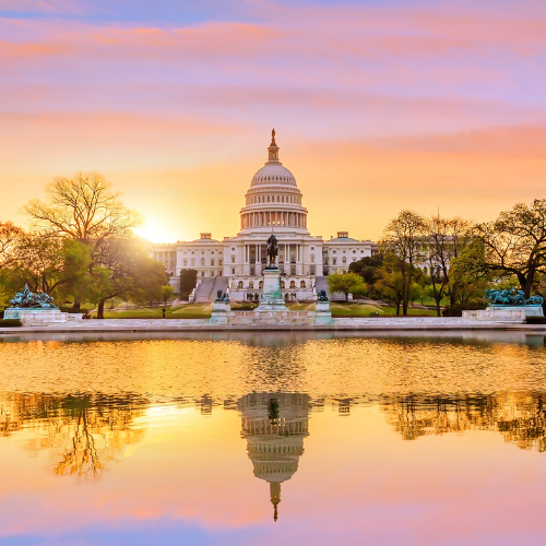U.S. Capitol building at sunset with warm orange sky, reflected in a calm pool in the foreground, surrounded by trees and a central statue.