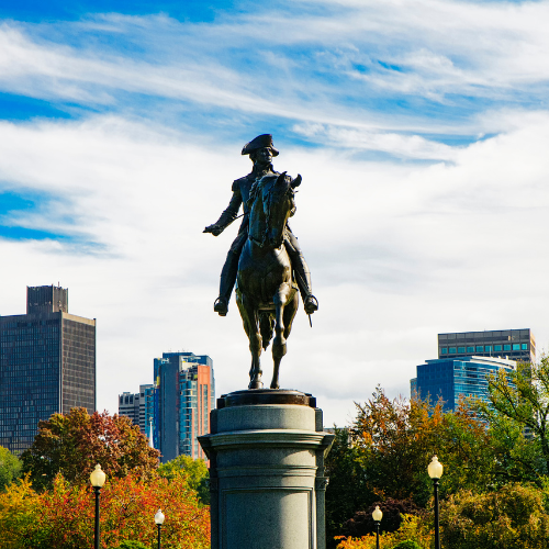 Equestrian statue of a historical figure in a city park with autumn trees and modern buildings in the background under a blue sky.