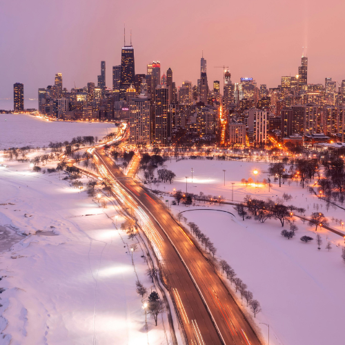 City skyline at dusk with illuminated buildings and a busy roadway leading into the city, bordered by a snow-covered park and waterfront.