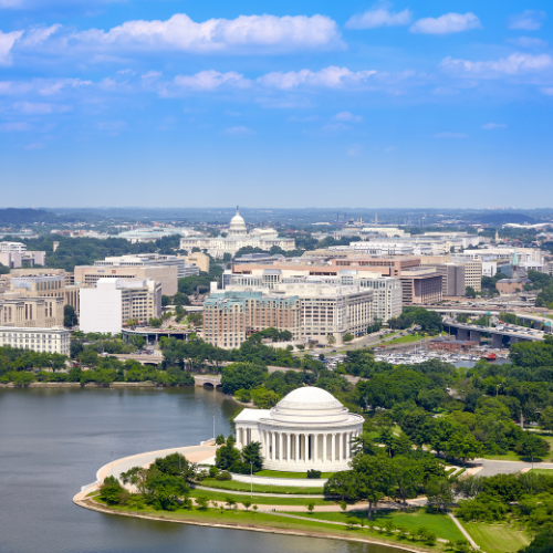 Aerial view of Washington, D.C. featuring the Jefferson Memorial in the foreground by the water and the U.S. Capitol in the distance under a blue sky.