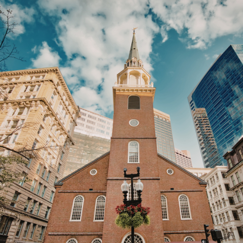 Historic brick church with a tall steeple surrounded by modern buildings under a partly cloudy sky