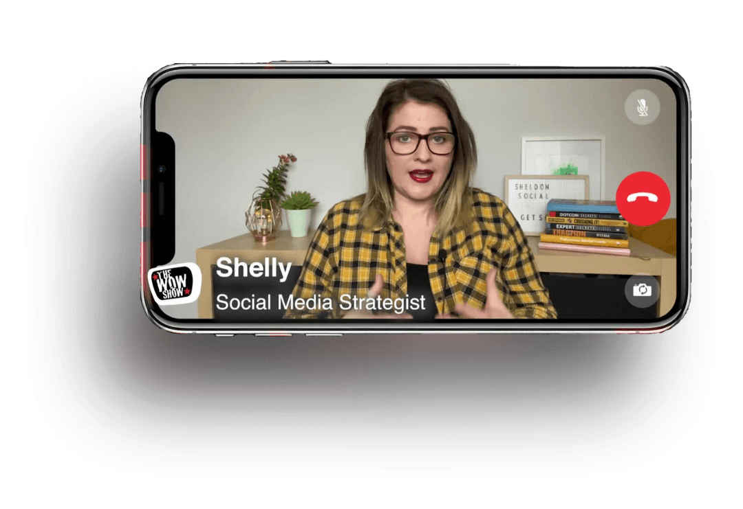 A woman with glasses and shoulder-length hair, wearing a yellow plaid shirt, appears on a smartphone screen during a video call. The background includes a desk with books, a small plant, and framed artwork.