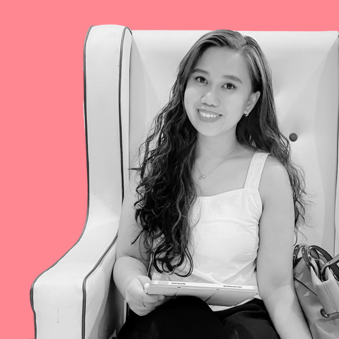 A young woman with long wavy hair, wearing a white sleeveless top, smiling and holding a tablet, sitting in a high-backed chair with a pink background.