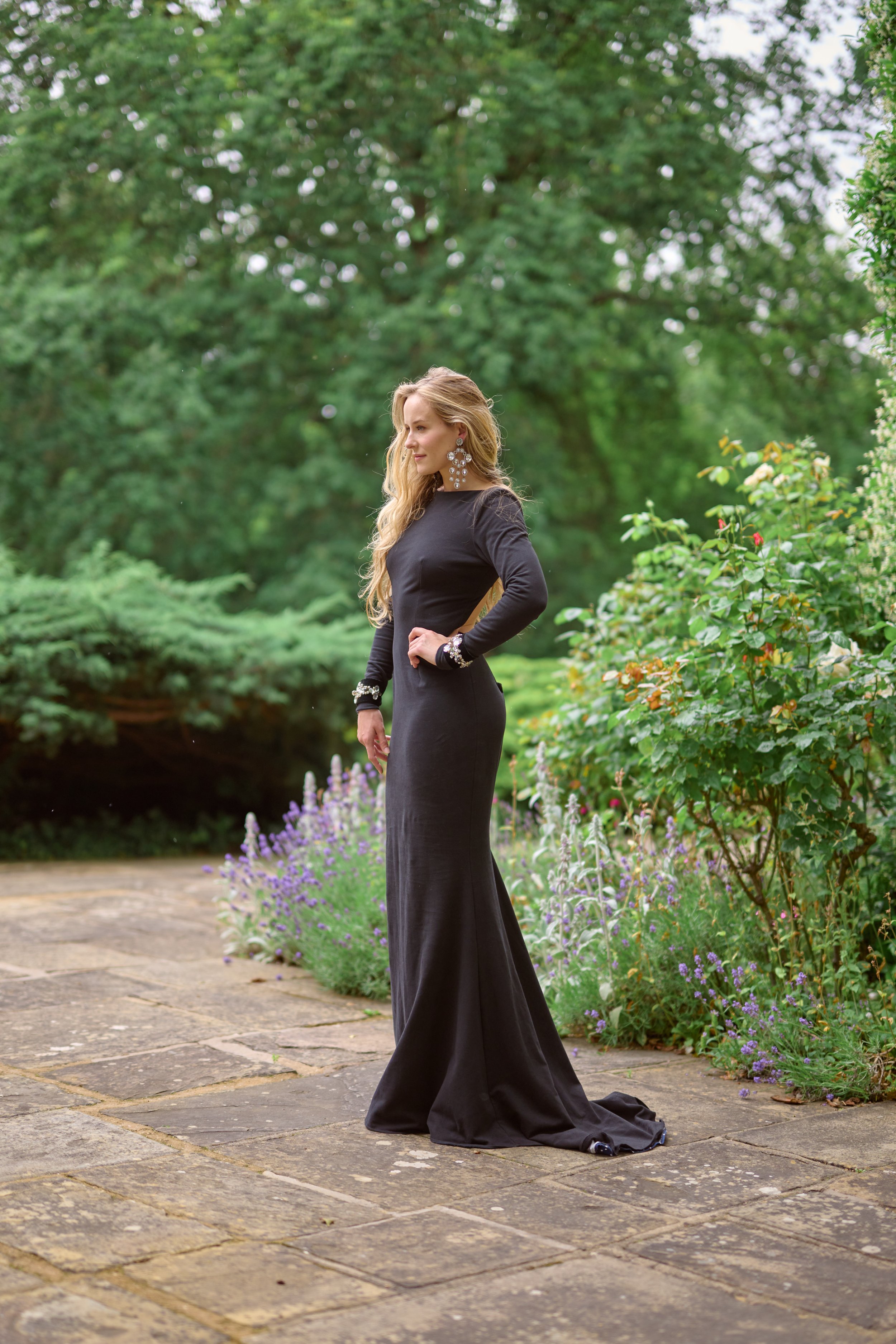 A woman in a black, long evening gown standing outdoors on a stone pathway surrounded by greenery and flowers.