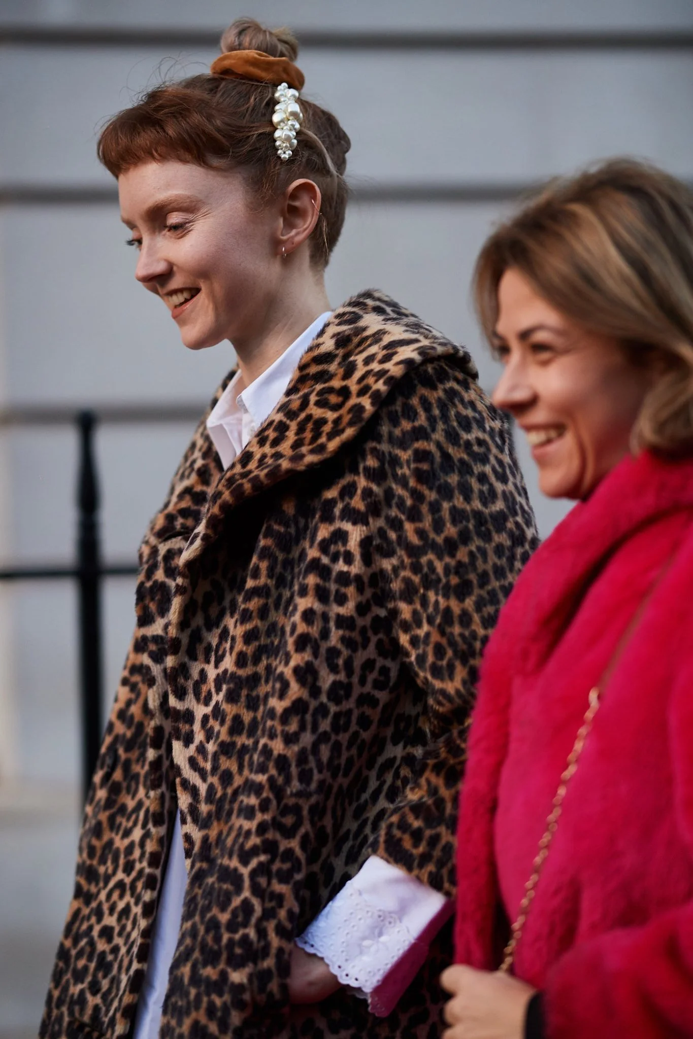 Two women walking together, smiling, one wearing a leopard print coat and the other in a bright pink coat, outdoors.
