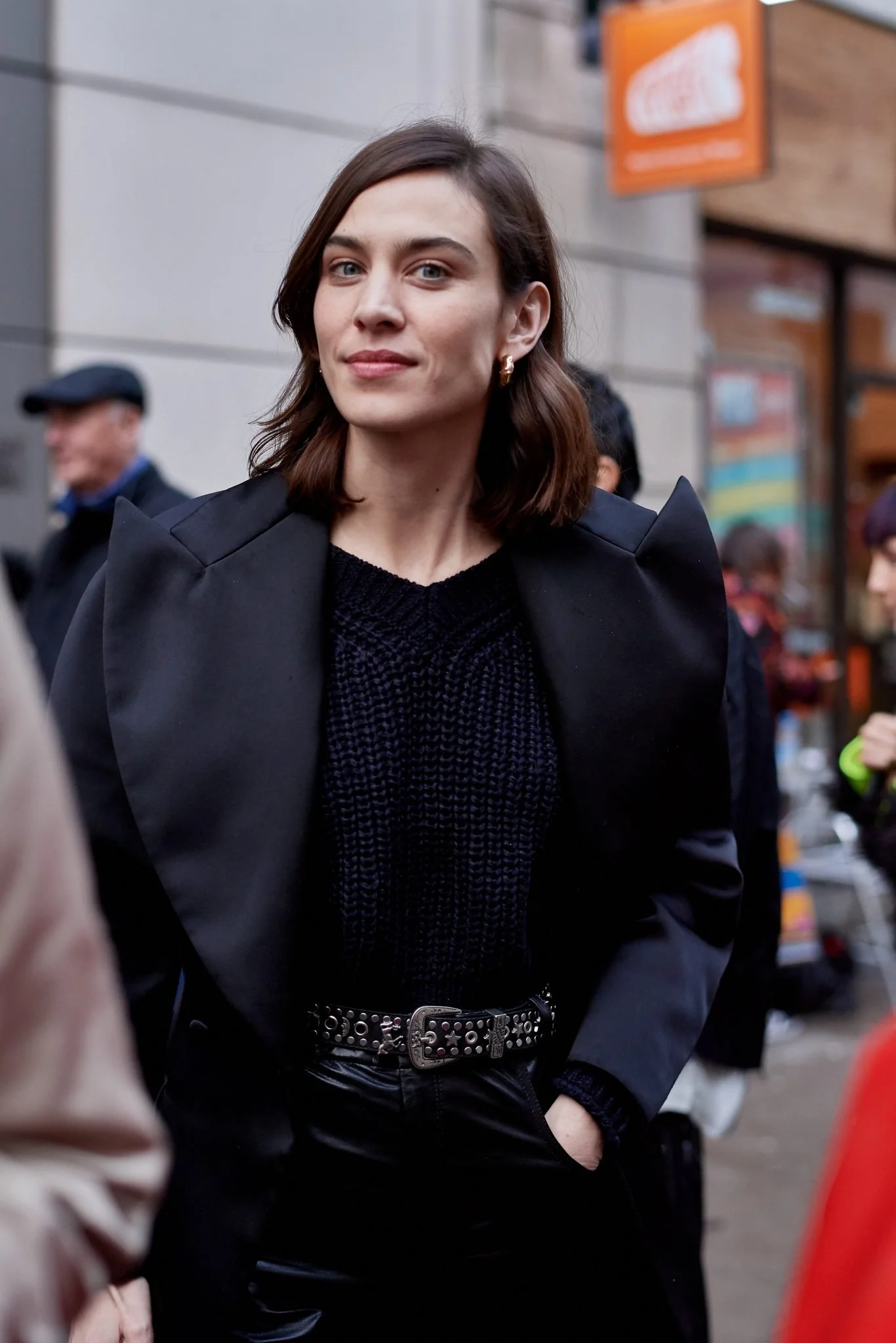 A woman with shoulder-length brown hair and light skin, wearing a black blazer and black sweater, standing outdoors in a city environment.