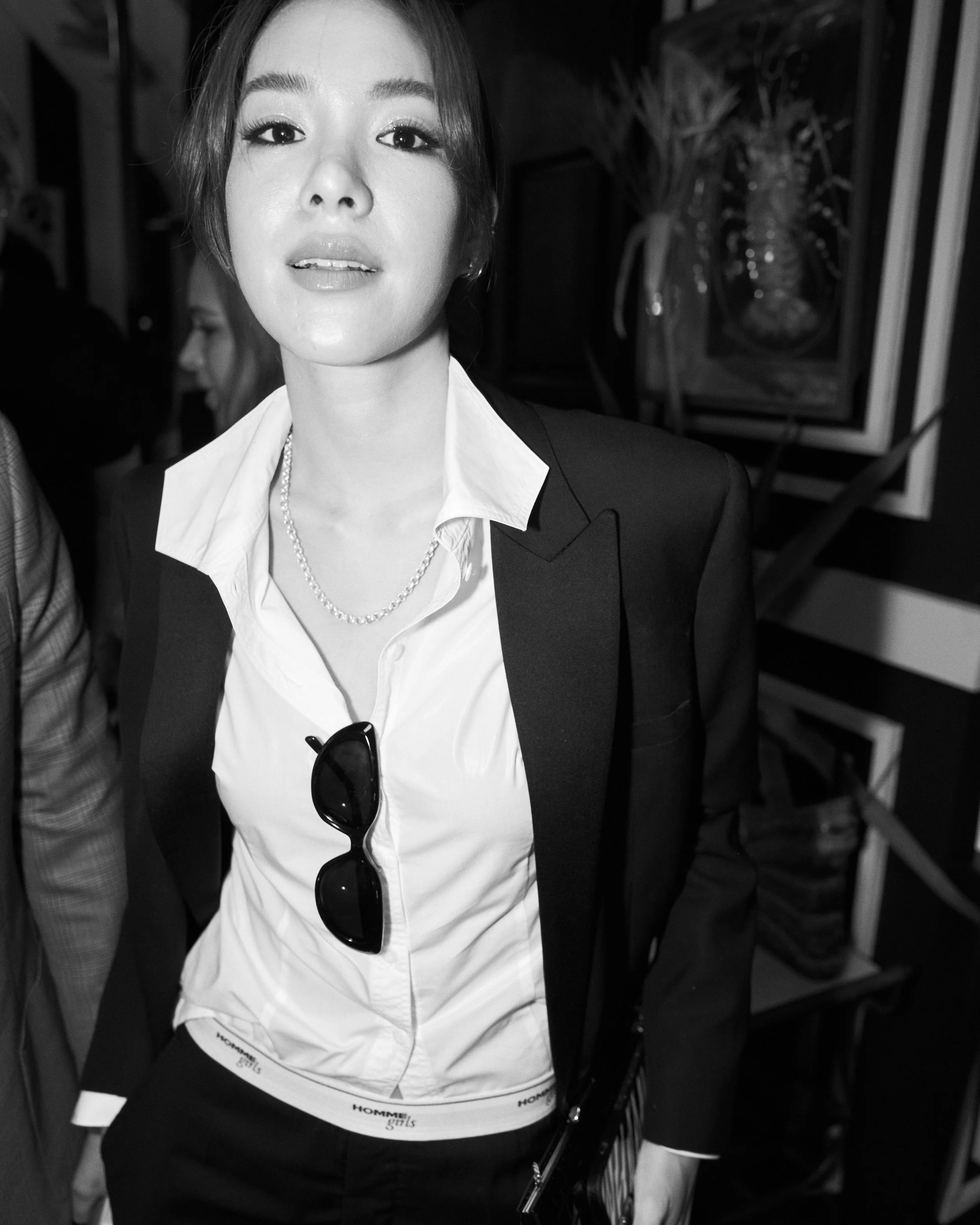 A woman in a black blazer and white shirt, wearing sunglasses hanging from her shirt and a pearl necklace, standing indoors with a blurred background including framed artwork and plants.