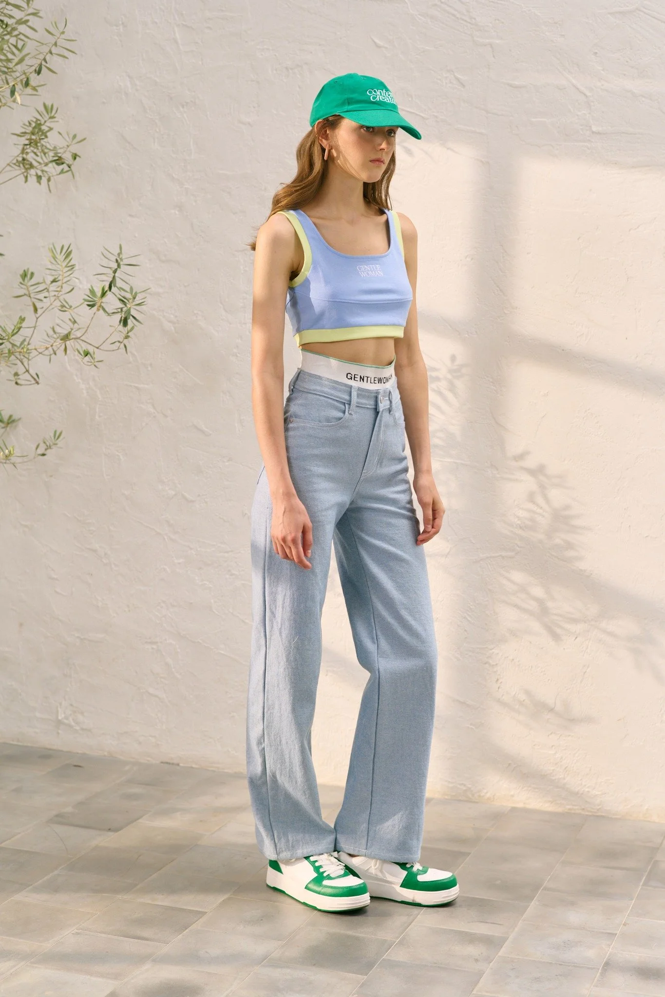 A young woman wearing a green cap, a pastel blue and yellow crop top, high-waisted light blue jeans, and green and white sneakers standing against a textured beige wall with a small plant on the left side.