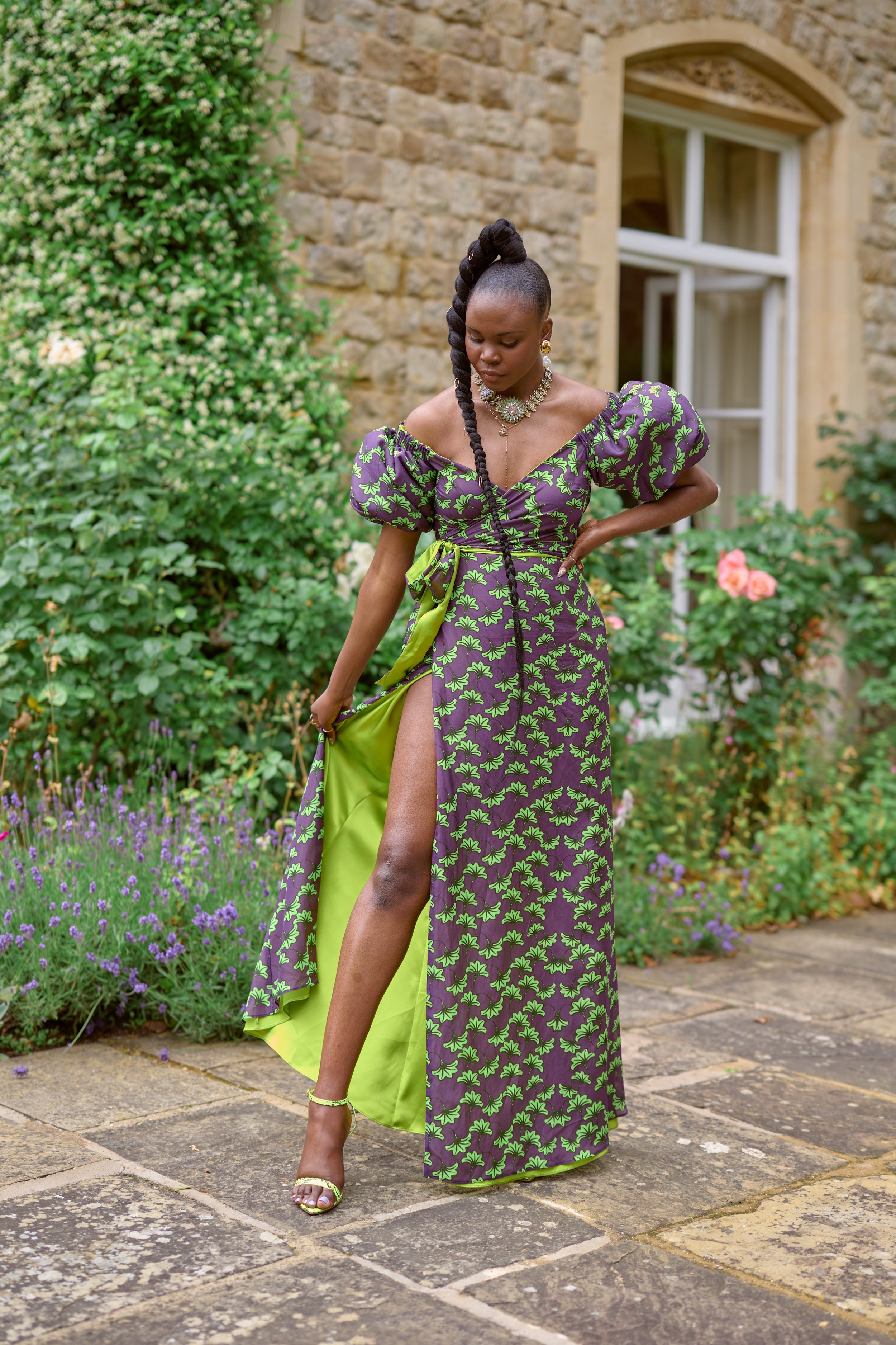A woman in a colorful dress with a high slit, standing outdoors on a stone pathway surrounded by green plants and flowers, with a stone house in the background.
