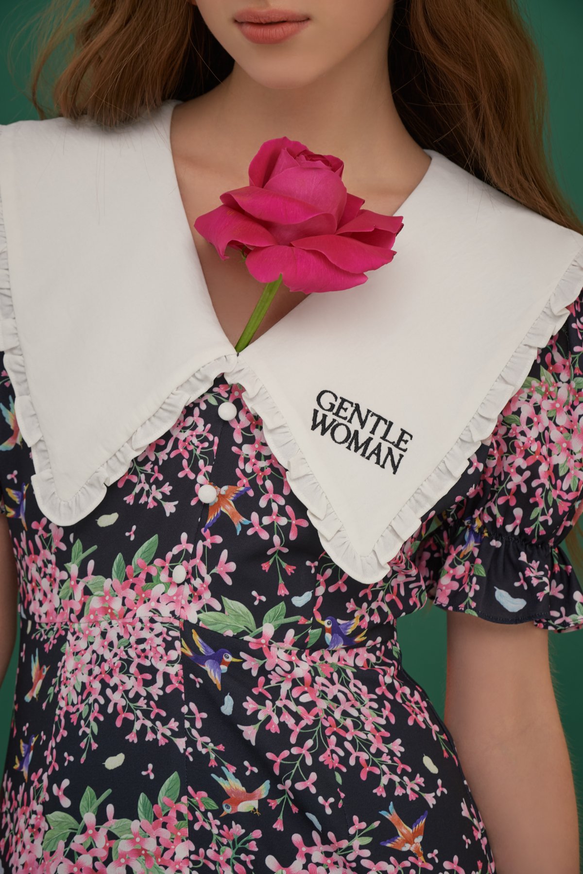 A woman wearing a floral dress with pink flowers and hummingbirds, a white collar with ruffled edges, and an embroidered label saying "Gentle Woman." She is holding a pink rose near her chest, with only the lower part of her face visible.