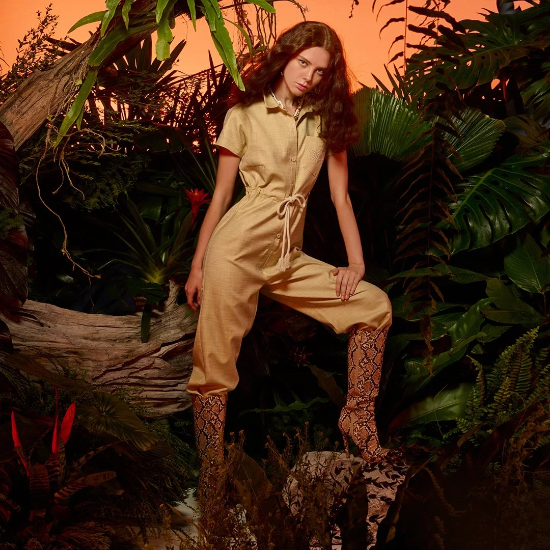 A woman with long curly hair wearing a beige jumpsuit and snake-print knee-high boots, standing in a lush tropical jungle setting with large green leaves and a glowing orange sunset background.
