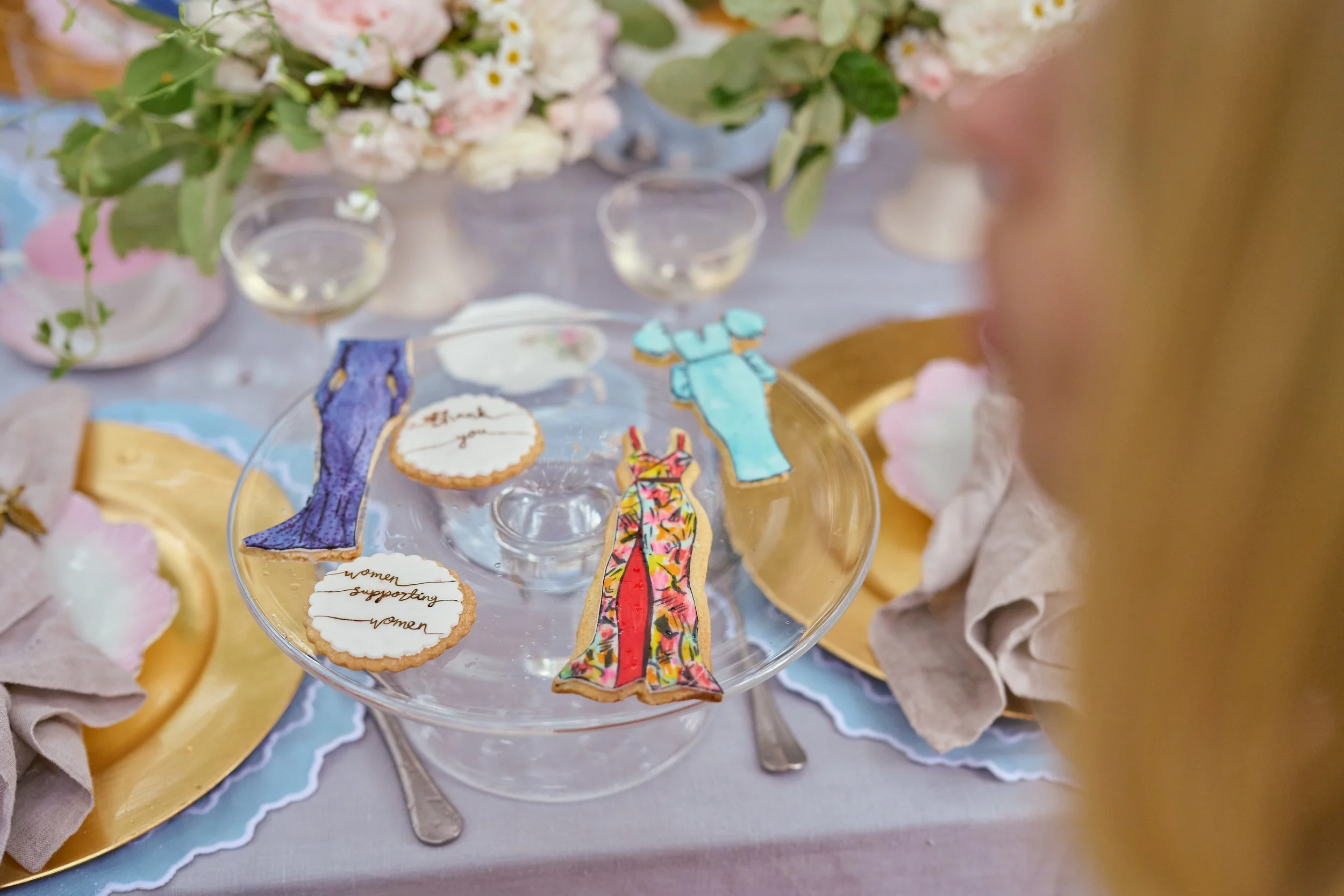 Decorated cookies on a glass stand with phrases supporting women, a bracelet, and a blue dress-shaped cookie, with floral arrangements and tableware in the background.