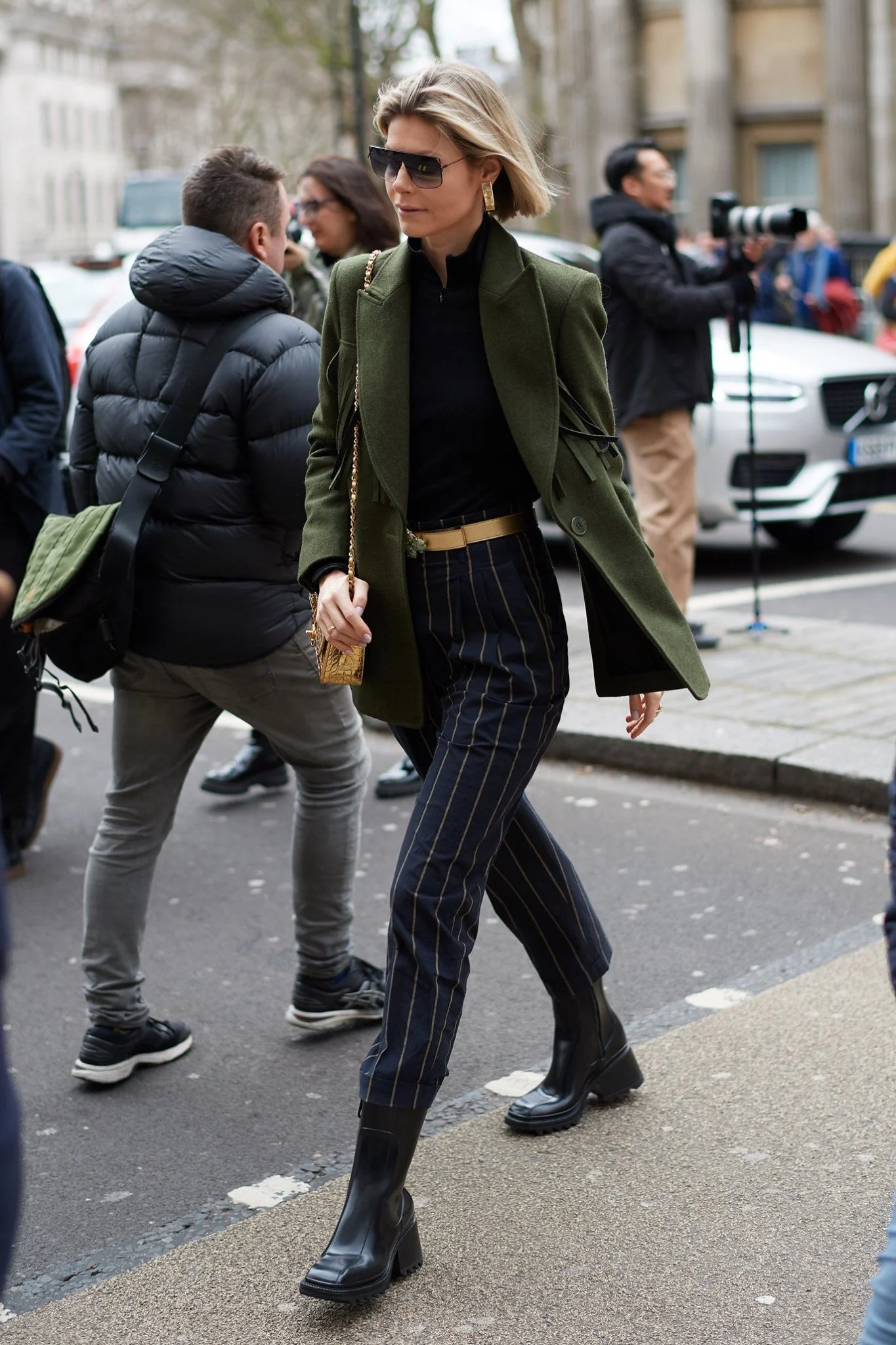A woman walking on a city street, wearing a green coat, black top, striped pants, black boots, sunglasses, and carrying a gold purse, with photographers and pedestrians in the background.