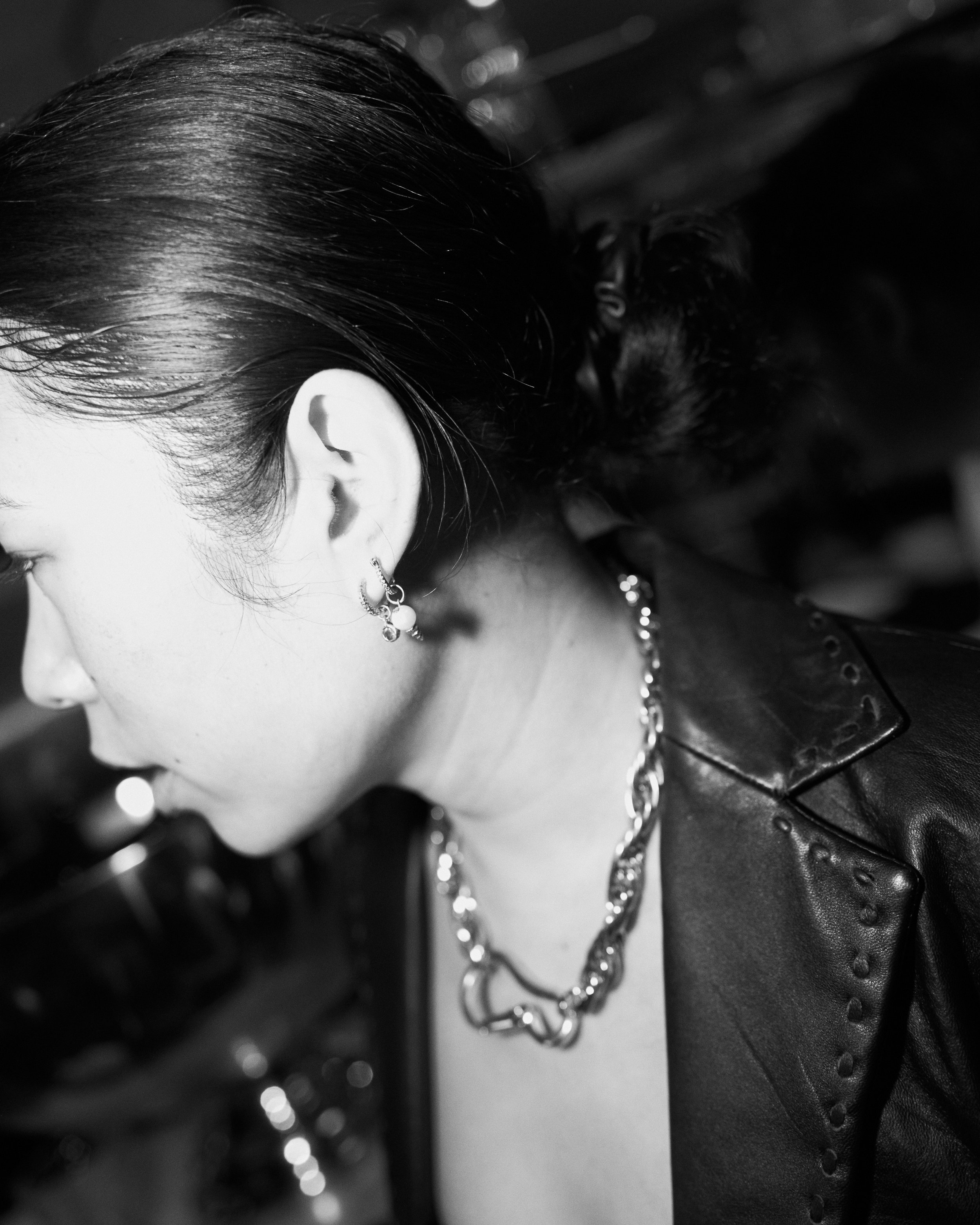 A woman with dark hair styled back, wearing earrings and a chunky chain necklace, dressed in a leather jacket, captured in black and white.