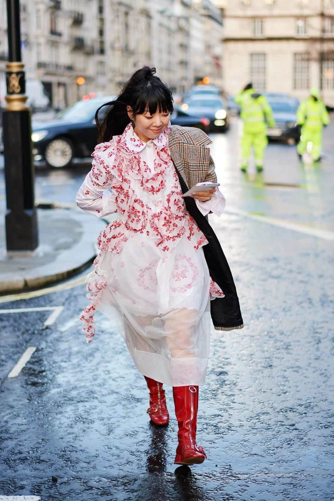 Woman wearing a white and red embroidered dress with red rain boots walking on wet city street, holding a phone and smiling, with city buildings and traffic in background.