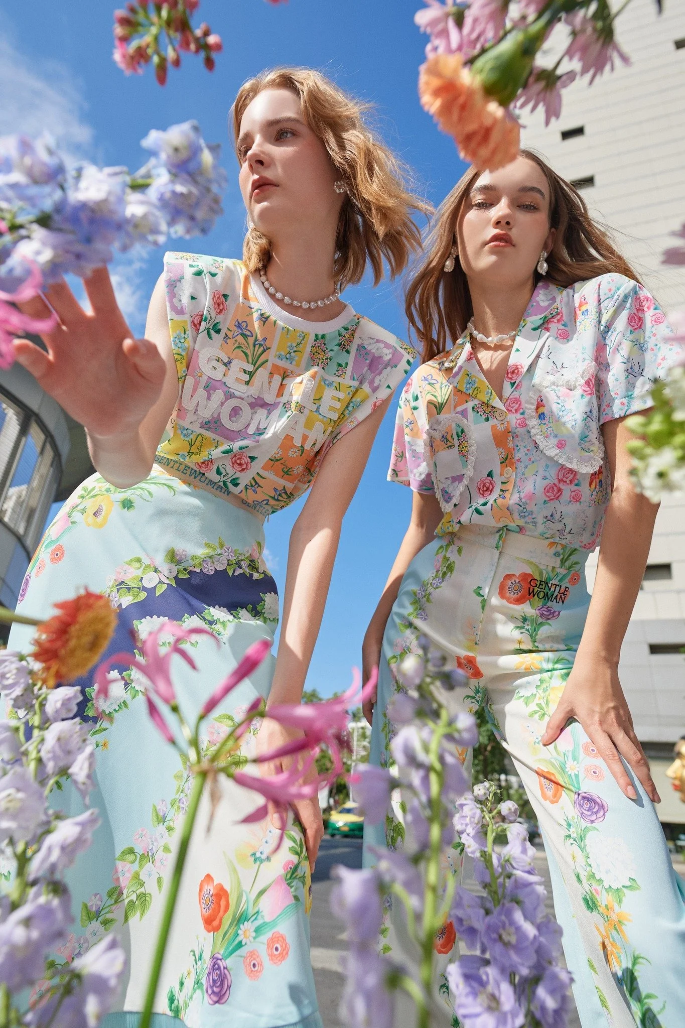 Two women standing outdoors surrounded by colorful flowers, wearing bright floral clothing with the words 'Gentle Woman' visible on their outfits, against a blue sky.