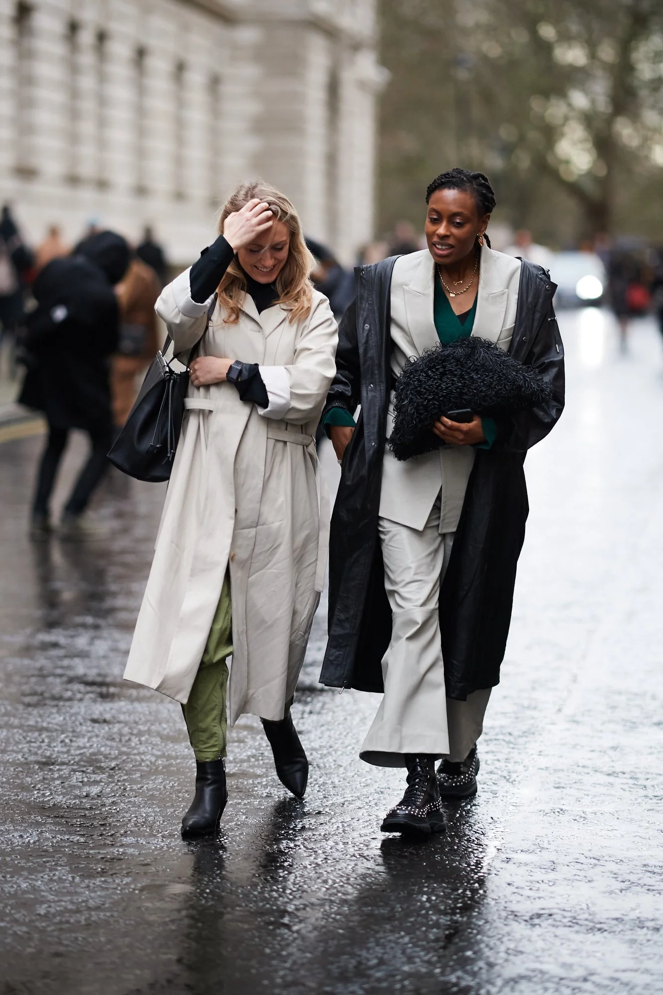 Two women walking together in rainy weather, dressed in stylish trench coats and boots, engaged in conversation.