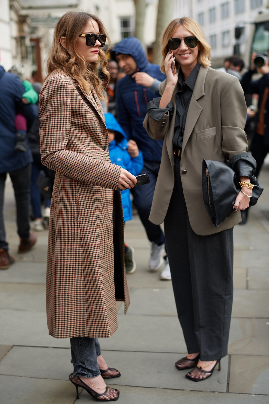 Two women standing outdoors on a city sidewalk, dressed in stylish outfits, wearing sunglasses, and talking on their phones, surrounded by other people.