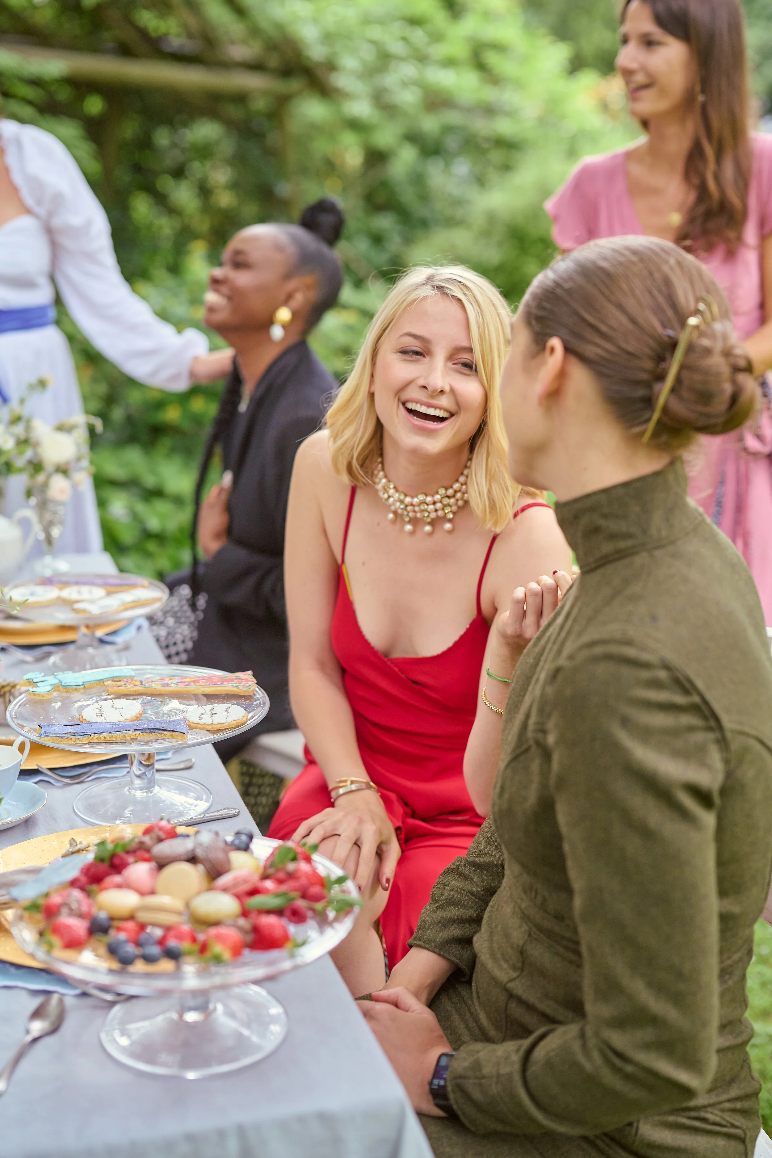 Women enjoying a gathering outdoors with a table of desserts and food, smiling and engaged in conversation, surrounded by greenery.