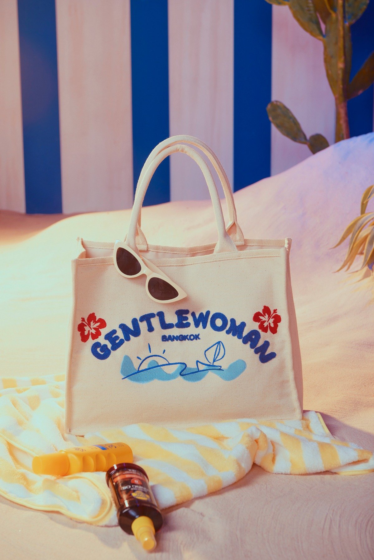 A tote bag with the words 'Gentlewoman Bangkok' and illustrations of a sun, sailboat, and waves. Sunglasses hang on the bag. Items like sunscreen and sunglasses are placed on a striped towel. A plant is in the background with a blue and white striped