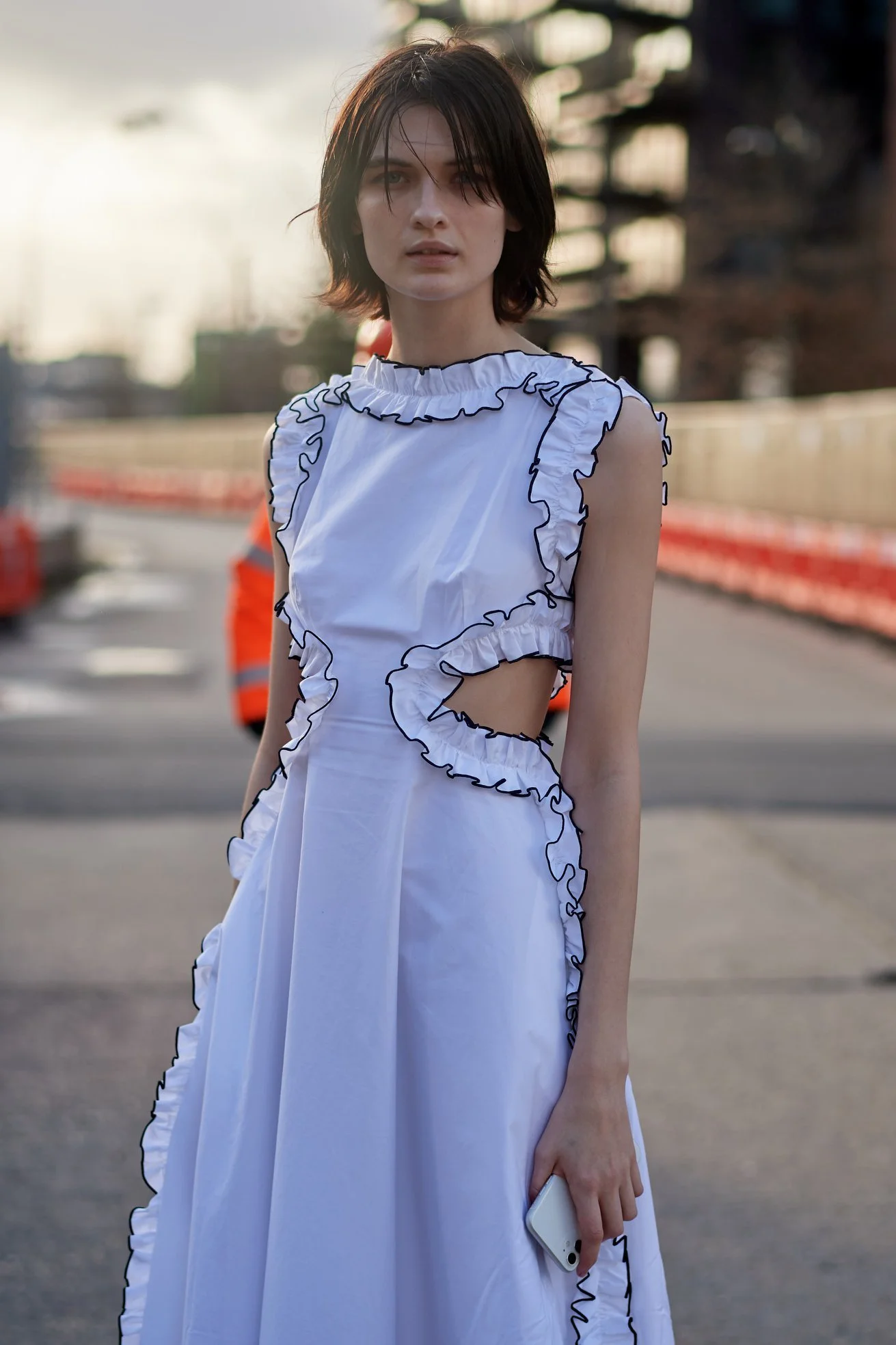 A young woman with short dark hair wearing a white dress with black ruffle trim, standing outdoors on a city street during daytime, holding a phone in her right hand.