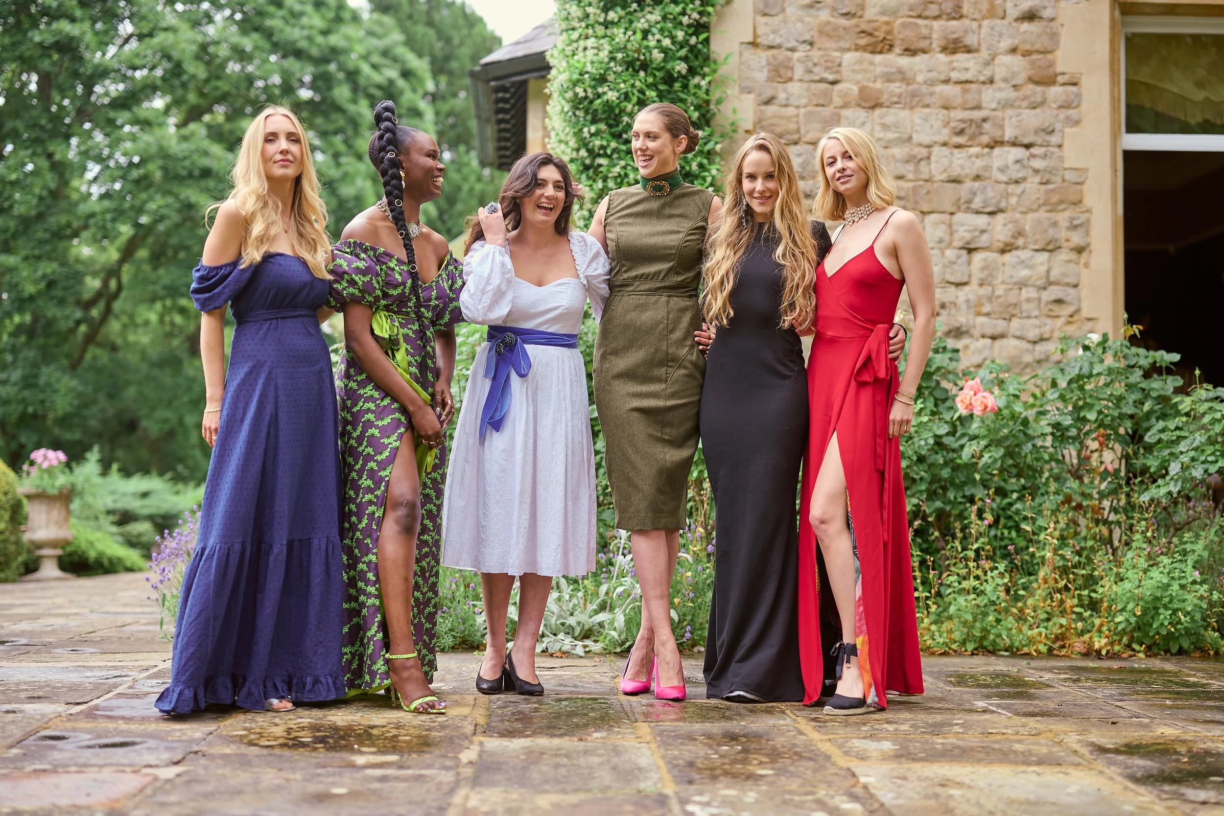Six women in colorful dresses standing together outdoors in a garden, smiling and posing for a photo.