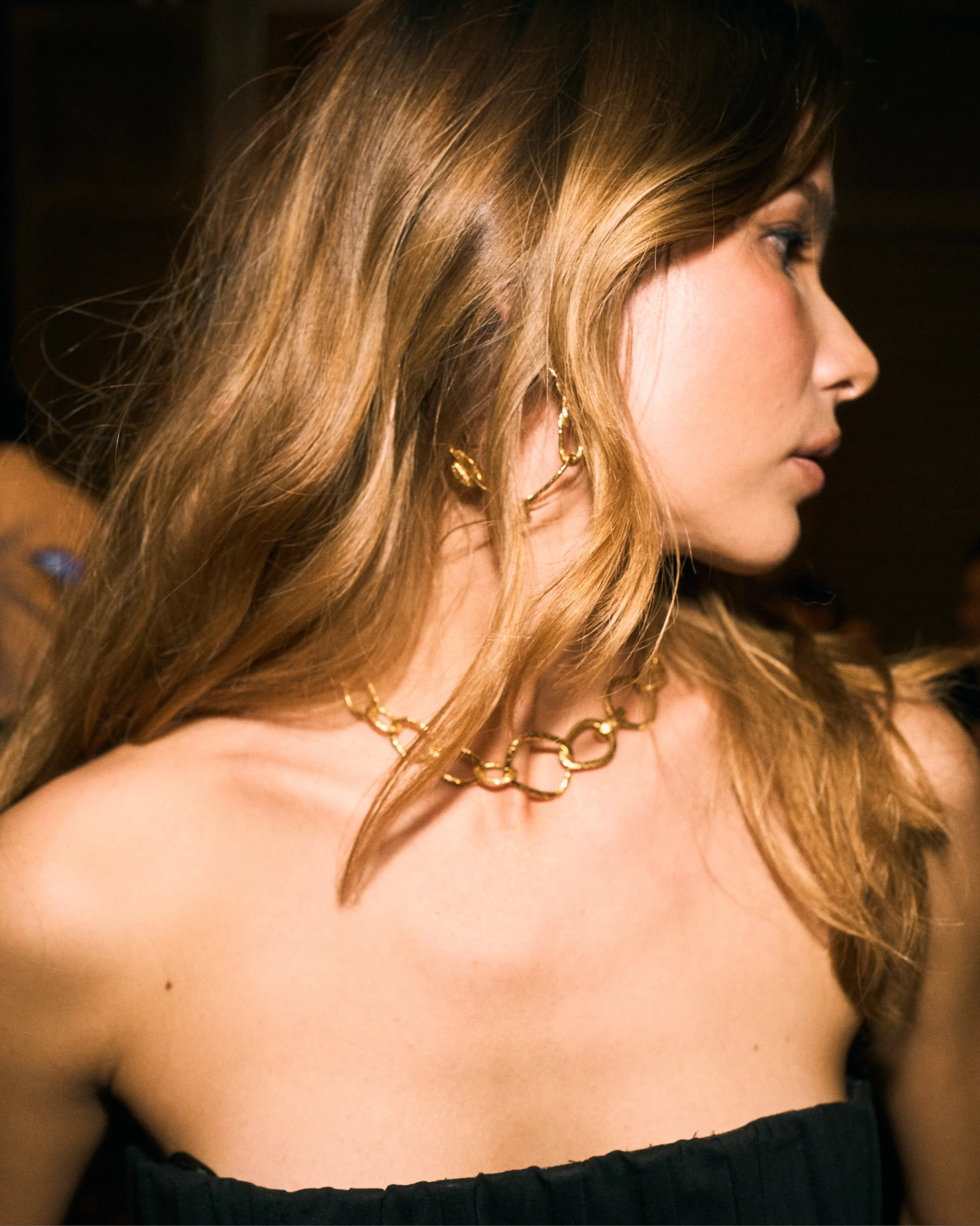 Profile of a woman with long, wavy blonde hair wearing gold jewelry, including a chain necklace, earrings, and a body chain, with a black strapless top.