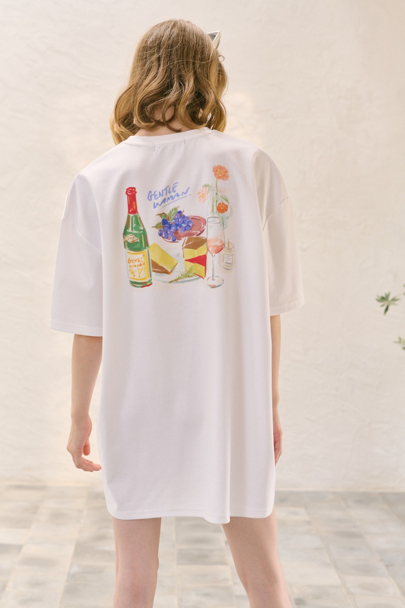 Back view of a person wearing a white T-shirt with colorful illustrations of wine, cheese, grapes, flowers, and the words "GENTLE WOMAN" on the back.