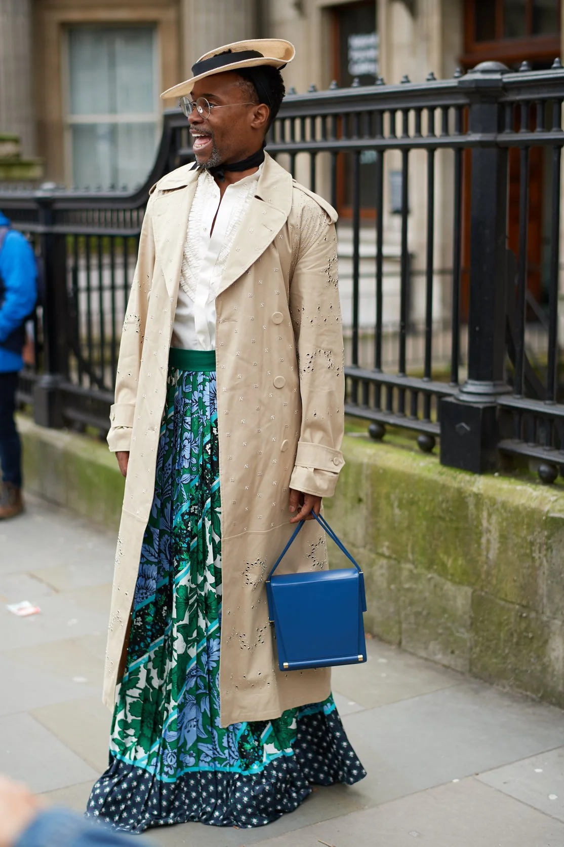 A person standing on a sidewalk wearing a beige trench coat, colorful wide-leg pants with floral patterns, a white shirt, a wide-brimmed hat, glasses, and carrying a blue handbag.