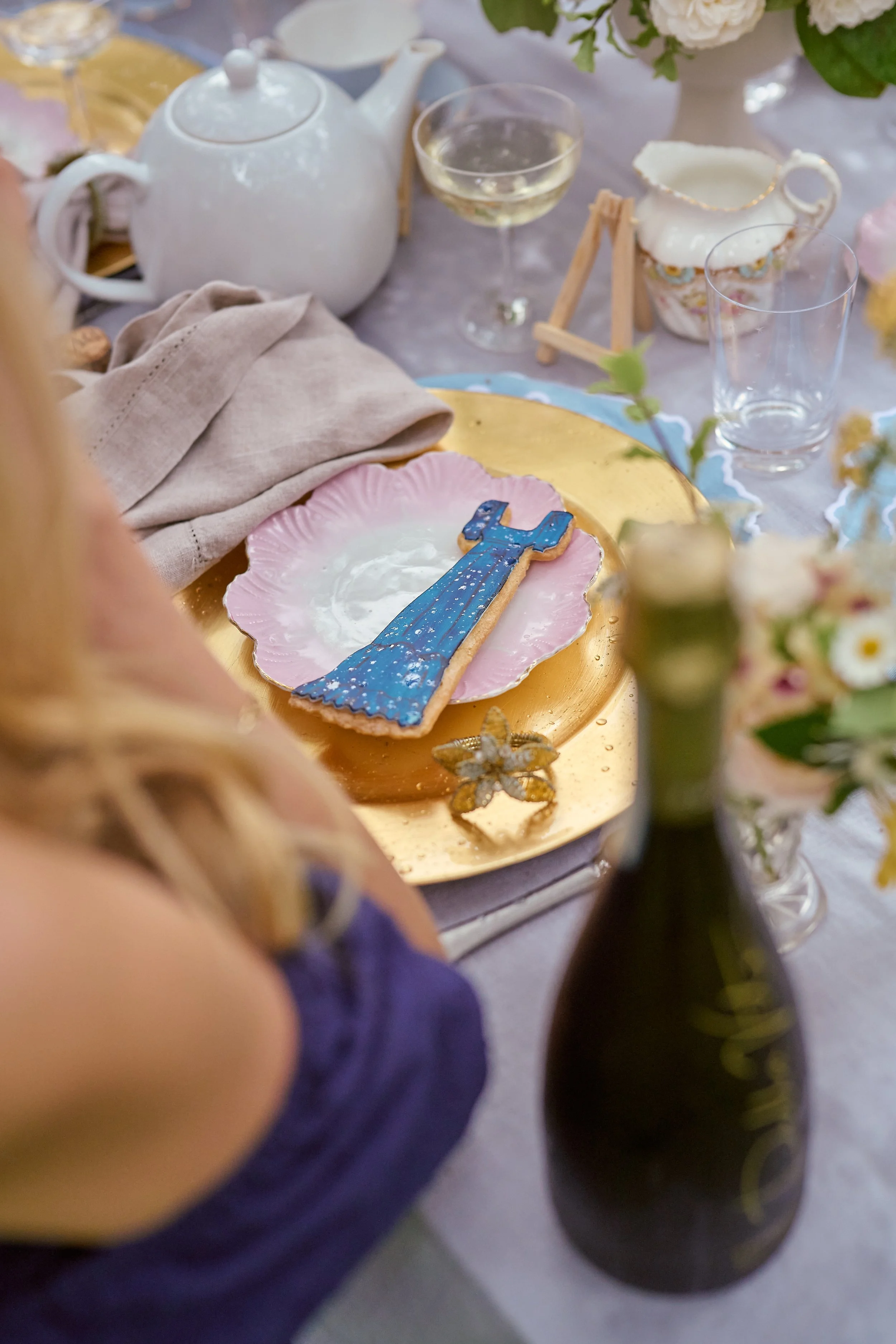 Table setting with a pink and gold plate holding a decorated sugar cookie shaped like a hammer, a teapot, wine glass, and floral arrangements.
