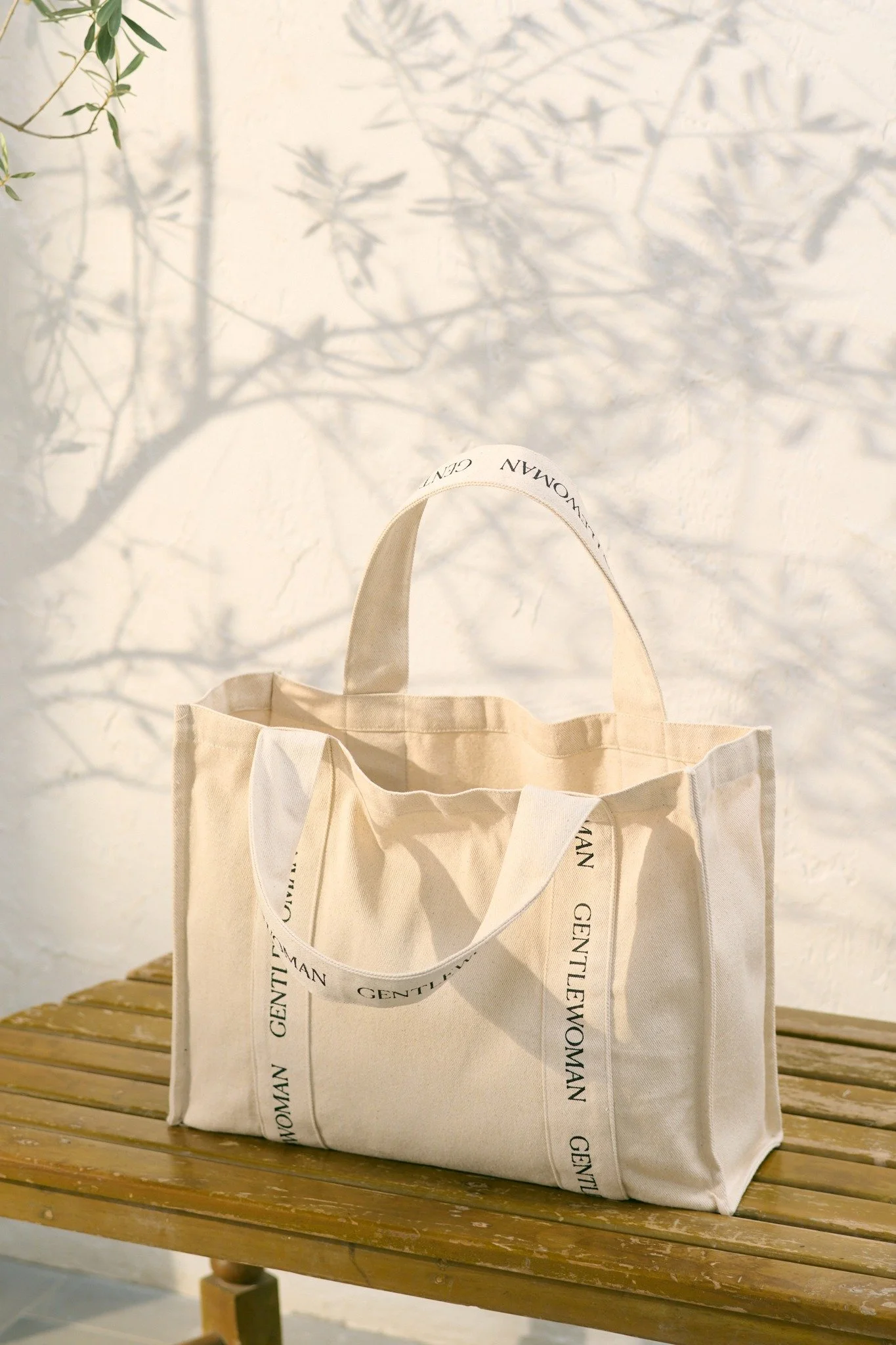 A cream-colored canvas tote bag with text 'GENTLEWOMAN' on the handles, placed on a wooden bench against a white wall with shadows of tree branches.