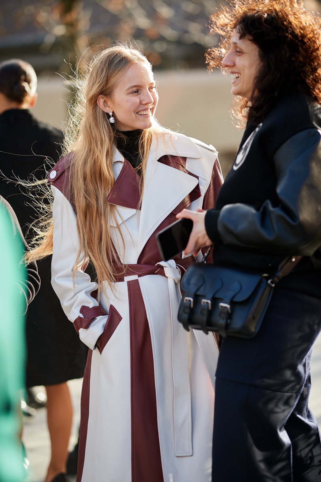 Two women, smiling and talking, outdoors during daytime. The woman on the left has long blonde hair and is wearing a beige and maroon coat. The woman on the right has curly hair and is dressed in dark clothing, holding a smartphone.