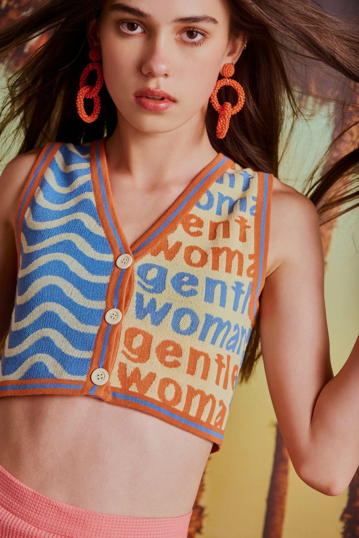 A young woman with long brown hair wearing large orange beaded earrings and a colorful sleeveless top with a mixed blue, orange, and cream pattern, standing in front of a tropical background with palm trees.