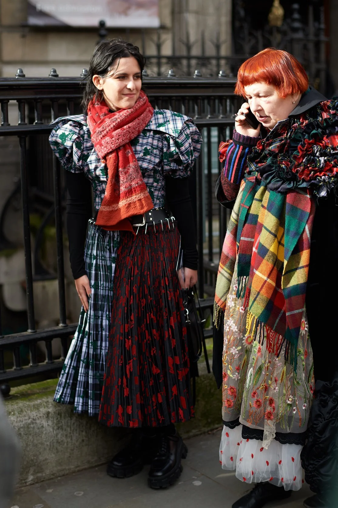 Two women dressed in eclectic, patterned clothing standing near a black fence outdoors. One woman has dark hair, a red scarf, plaid dress with ruffled sleeves, belt, long skirt, and platform boots. The other woman has red hair, floral-patterned scarf