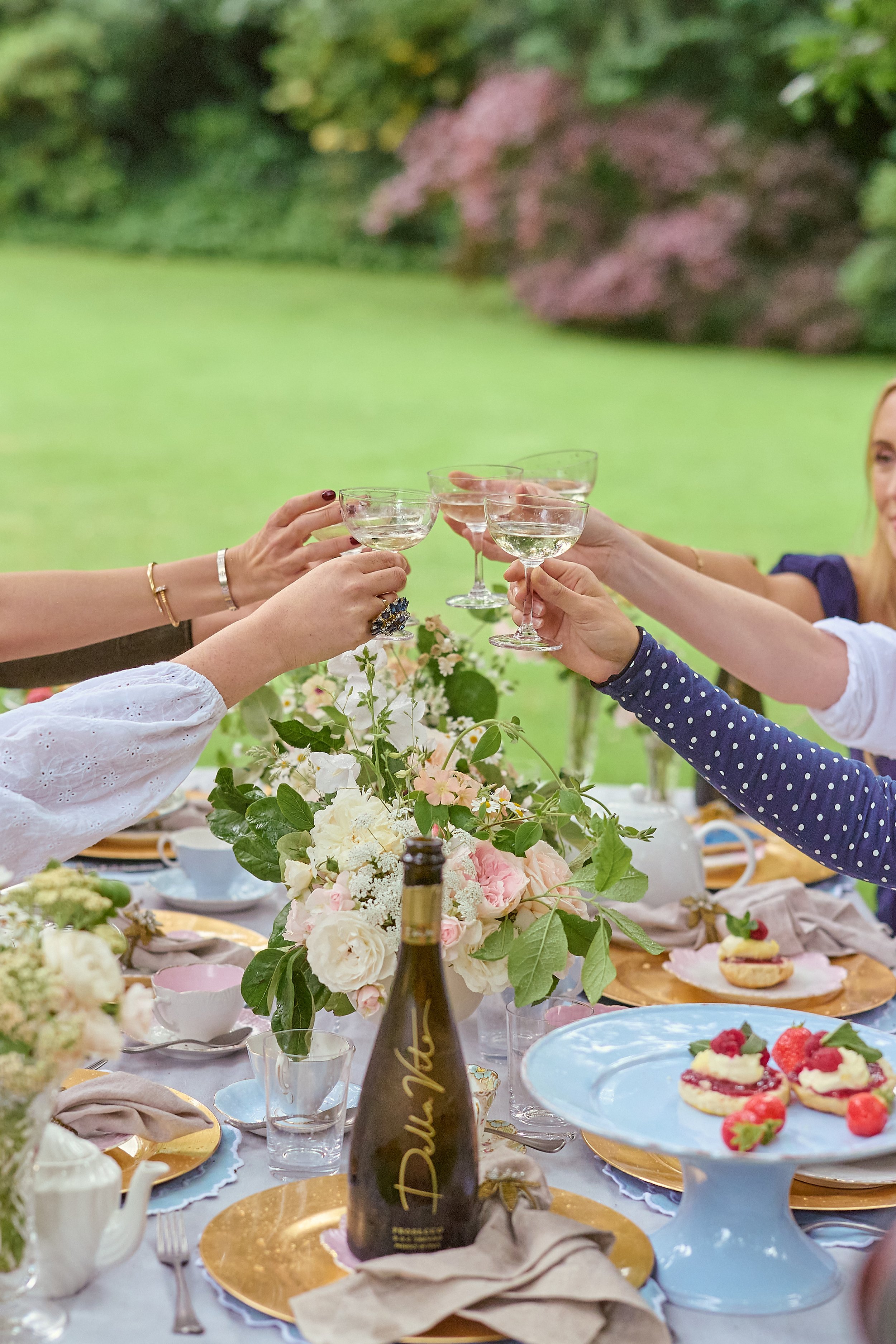 People raising glasses in a toast at an outdoor garden party with a decorated table, flowers, and desserts.