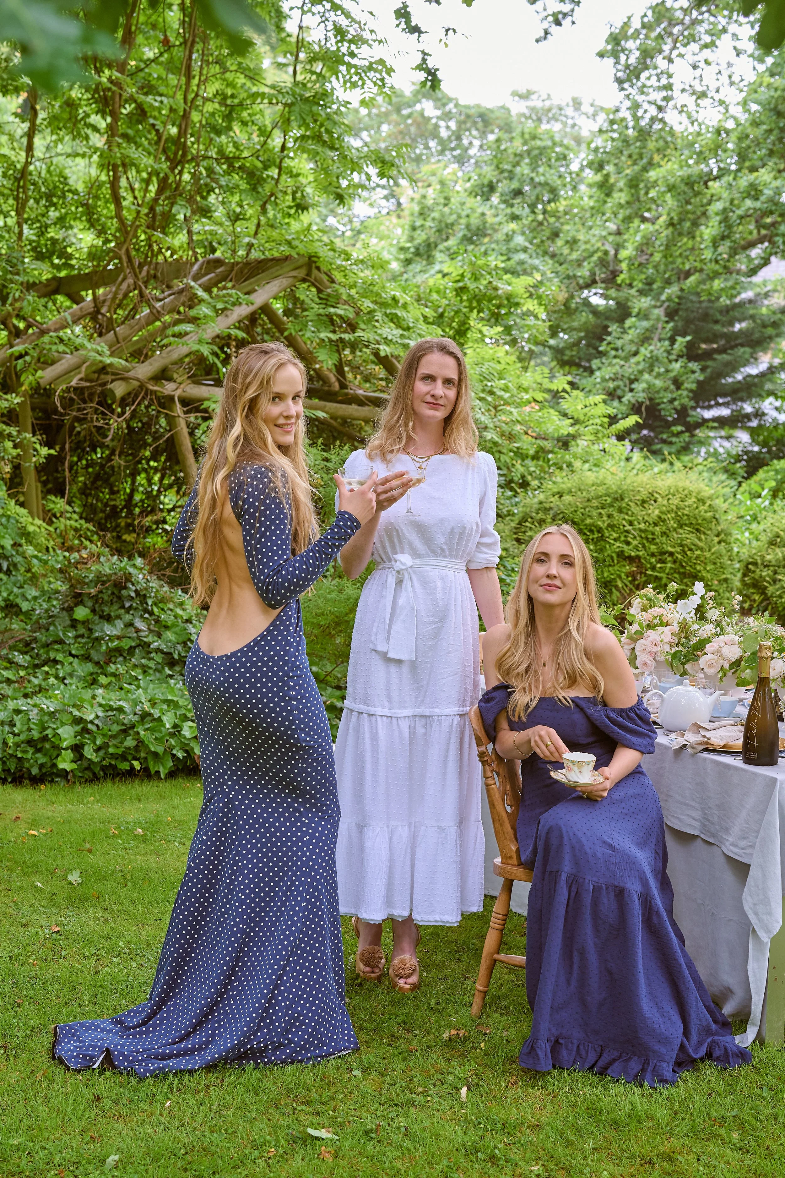 Three women in elegant dresses at a garden tea party, with a table set with flowers and tea cups.