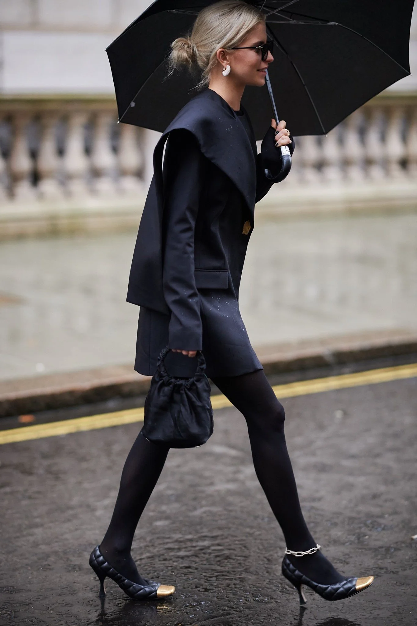 A stylish woman walking on a rainy day, holding a black umbrella, wearing black heels with gold accents, black stockings, a black skirt, a black blazer, and carrying a small black bag.