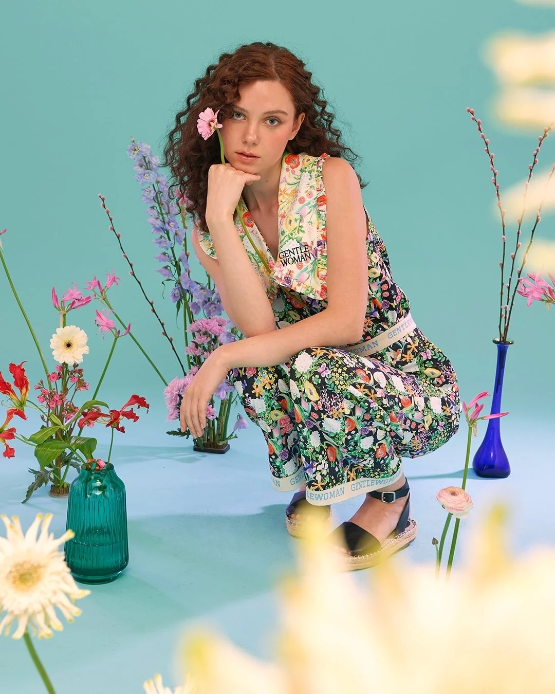 A young woman with curly red hair, sitting amid colorful flowers in vases, wearing a floral outfit with the words 'Gentle Woman' on the collar and waistband, against a pastel green background.