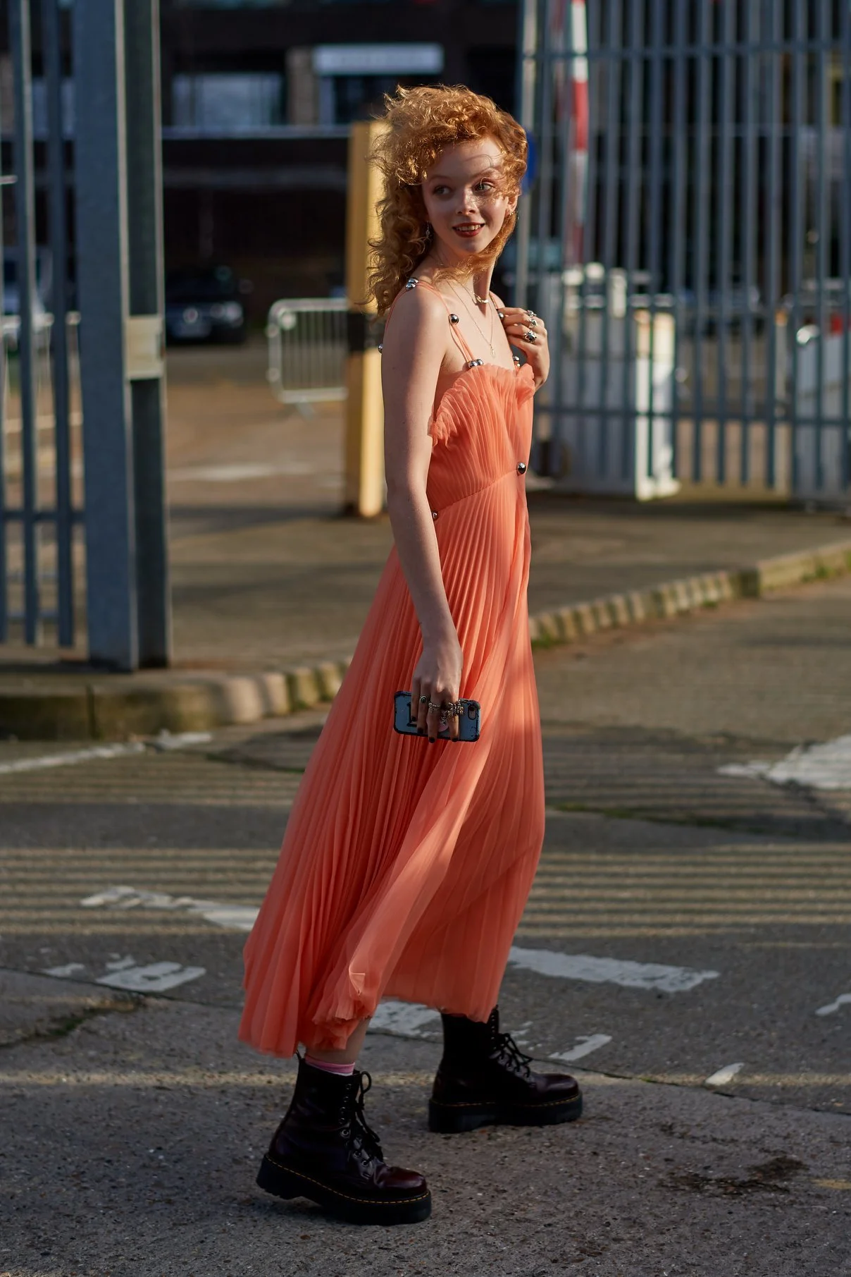 A woman with curly red hair wearing a peach-colored, pleated, sleeveless dress and black combat boots walking outdoors. She holds a smartphone in her right hand and is looking over her shoulder with a smile. The background features metal fencing and 